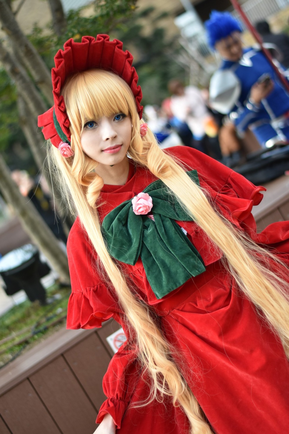 1girl blonde_hair blue_eyes blurry blurry_background blurry_foreground bonnet bow capelet depth_of_field dress lips long_hair long_sleeves looking_at_viewer multiple_girls photo realistic red_dress shinku solo solo_focus very_long_hair