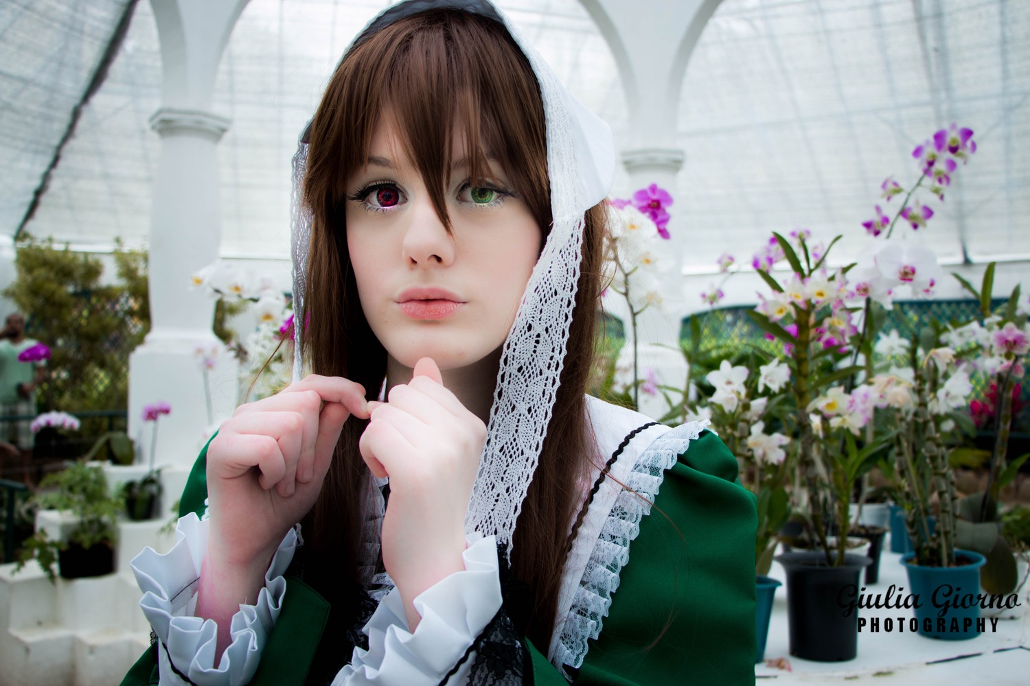 1girl 3d blurry brown_hair building closed_mouth depth_of_field dress fence flower frills green_dress green_eyes heterochromia lips long_hair long_sleeves looking_at_viewer outdoors plant red_eyes solo suiseiseki upper_body