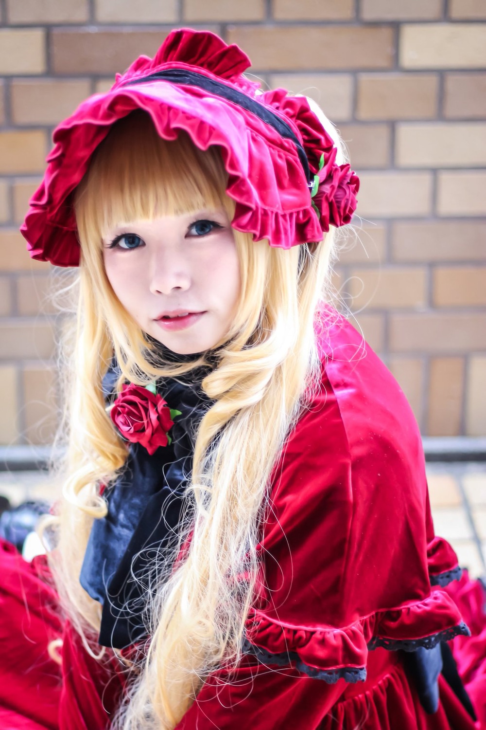 1girl bangs blonde_hair blue_eyes blurry blurry_background bonnet building capelet depth_of_field dress flower lips long_hair looking_at_viewer photo red_capelet red_dress red_flower red_rose rose shinku solo upper_body