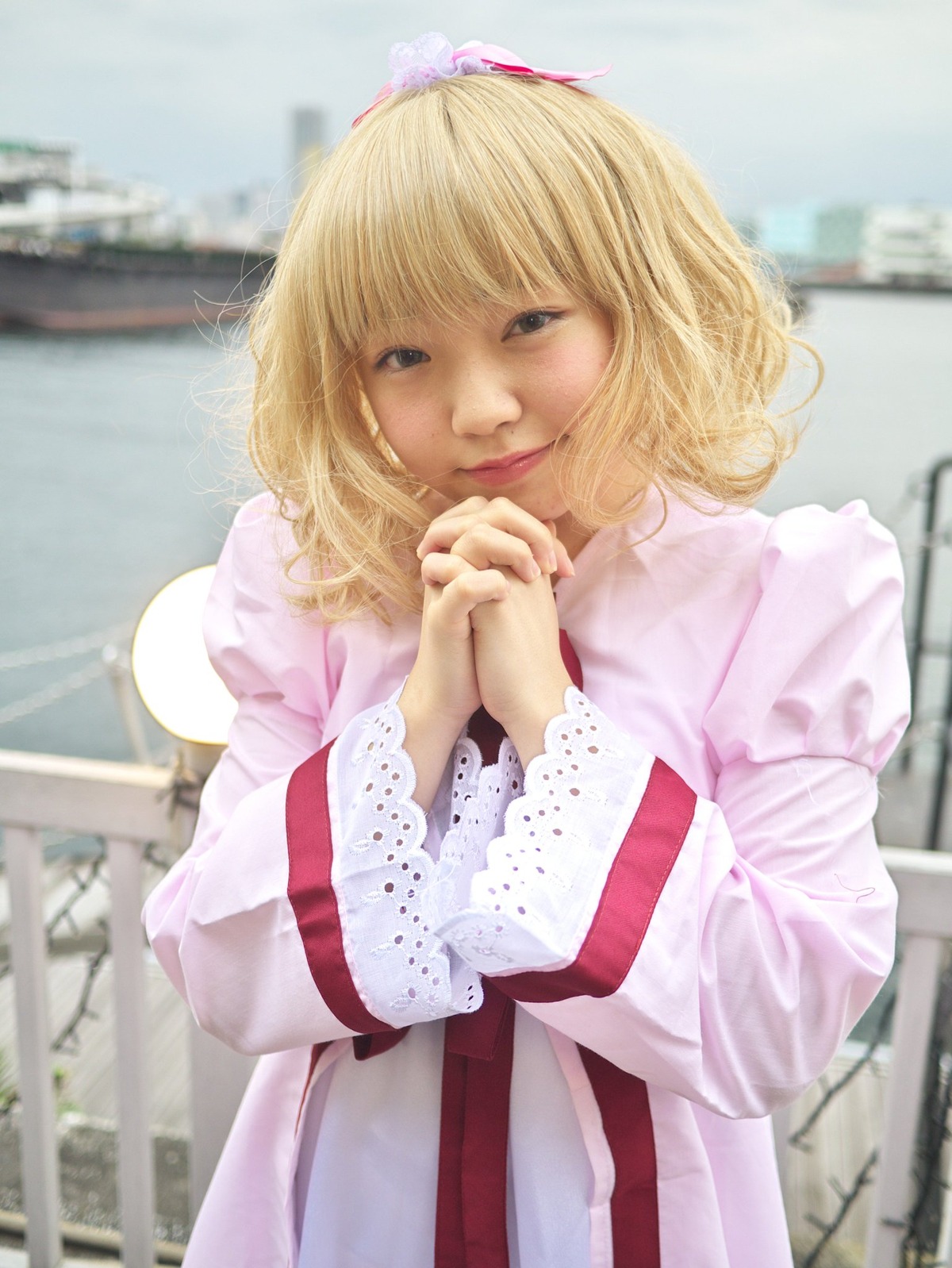 1girl alice_margatroid bangs blonde_hair blue_eyes blurry blurry_background building city day depth_of_field dress hairband hands_clasped hands_together hinaichigo lips long_sleeves looking_at_viewer outdoors own_hands_together photo railing realistic short_hair sky solo upper_body