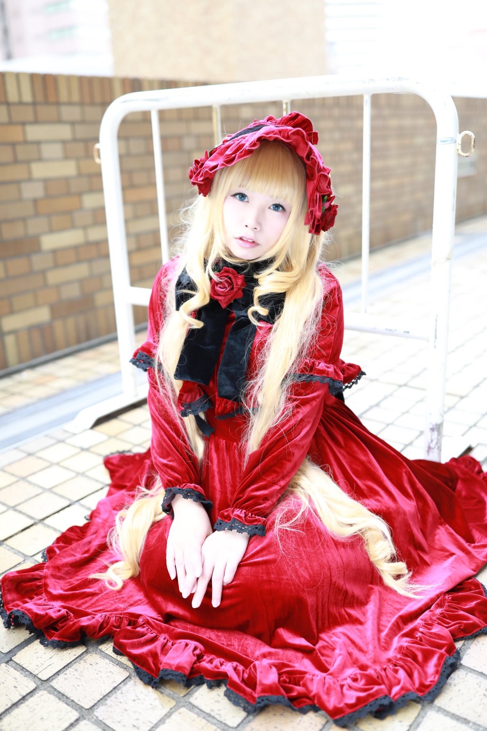 1girl blonde_hair blue_eyes blurry bonnet building depth_of_field dress flower frills long_hair long_sleeves looking_at_viewer photo red_dress rose shinku sitting solo tile_floor tiles