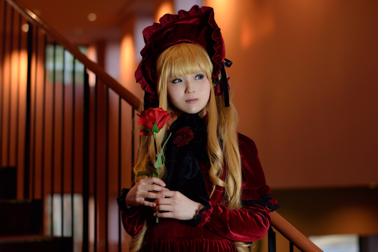 1girl bangs blonde_hair blue_eyes blurry blurry_background bonnet bookshelf depth_of_field dress flower lips long_hair long_sleeves red_dress red_flower red_rose rose shinku solo table