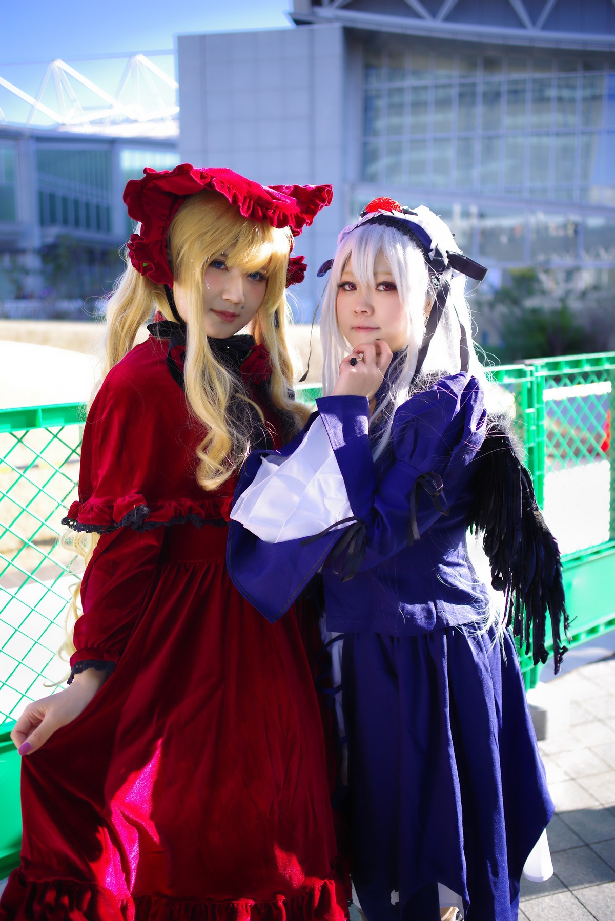 2girls blonde_hair blue_eyes blurry bonnet building capelet chain-link_fence city depth_of_field dress fence long_hair long_sleeves looking_at_viewer multiple_cosplay multiple_girls red_dress shinku stained_glass standing tagme tile_floor tiles