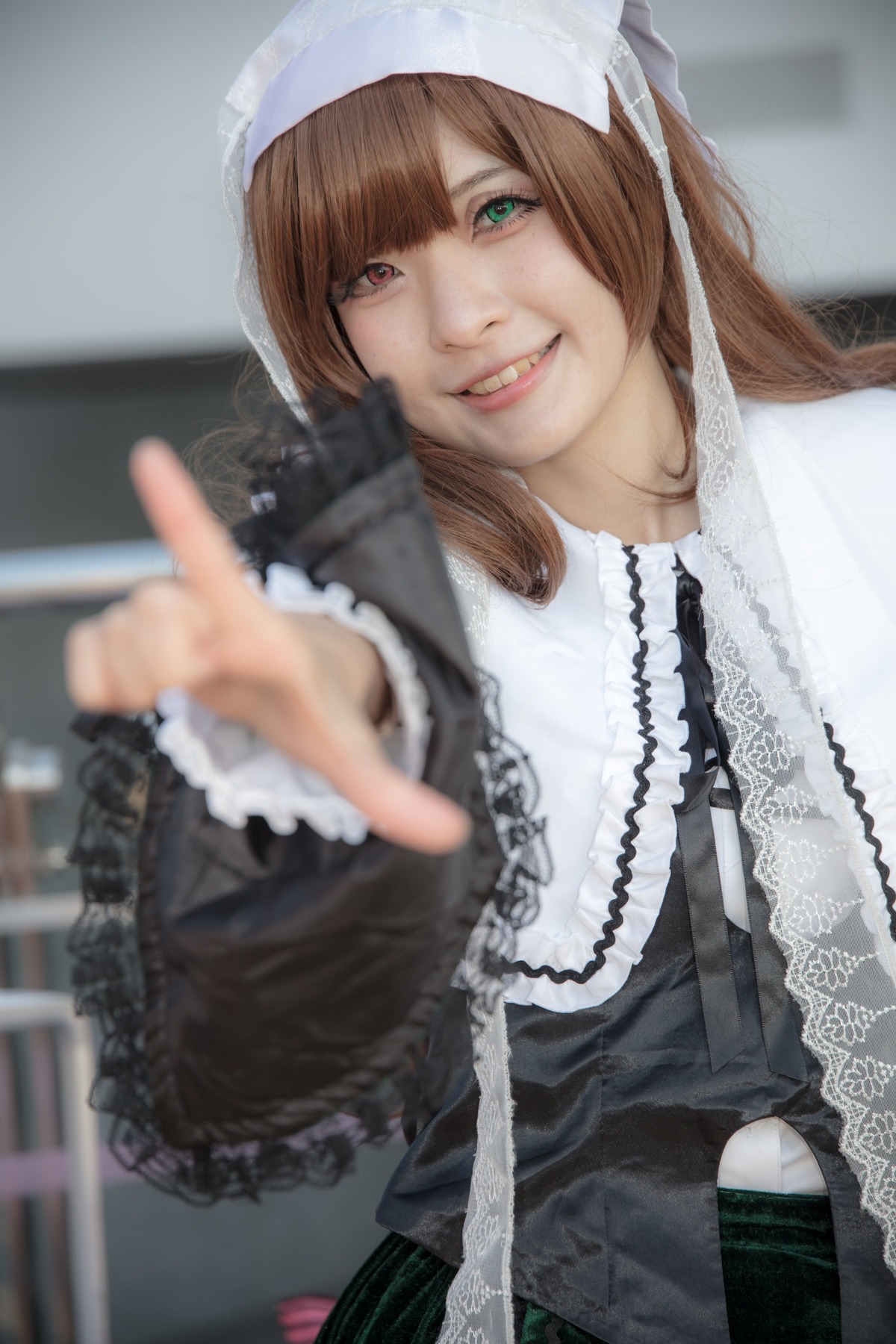 1girl 3d blurry blurry_background blurry_foreground brown_hair depth_of_field dress green_eyes grin heterochromia index_finger_raised lips lolita_fashion long_hair looking_at_viewer outstretched_arm photo pointing pointing_at_viewer reaching_out red_eyes smile solo suiseiseki