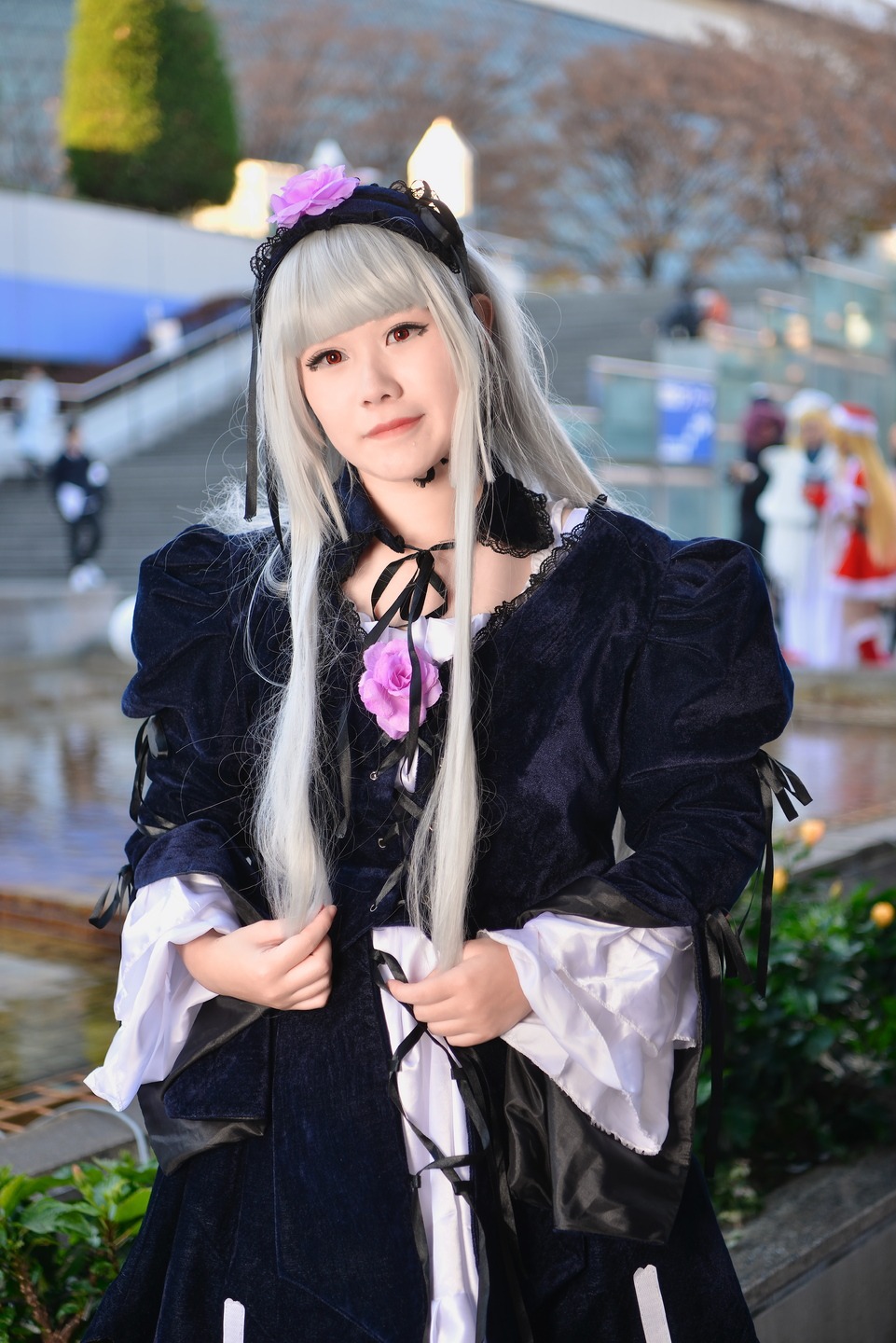 1girl black_dress blurry blurry_background blurry_foreground brown_eyes depth_of_field dress flower gothic_lolita hairband lips long_hair long_sleeves looking_at_viewer outdoors photo ribbon solo solo_focus standing suigintou